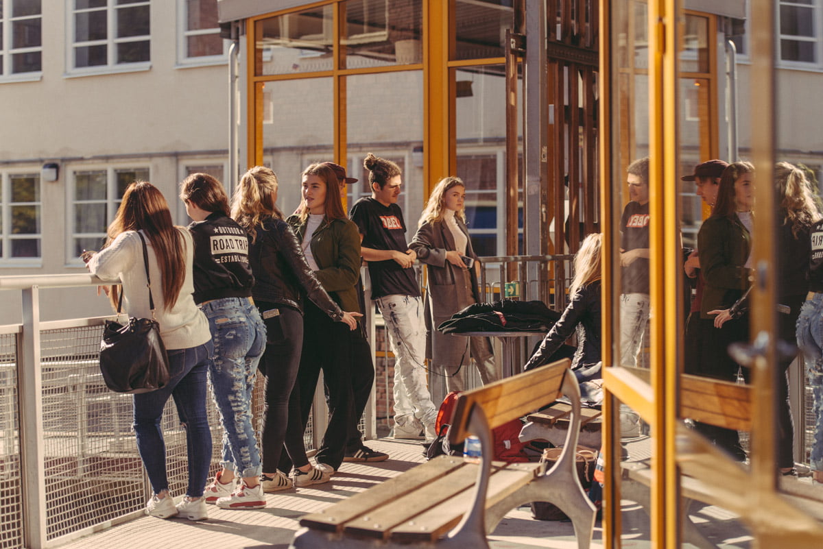 En grupp elever står och sitter i samtal på en solig skolgård vid en byggnad med stora fönster.