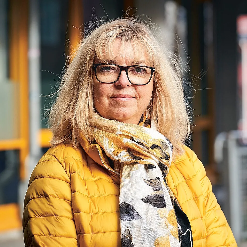SYV Katarina Karlquist står utomhuis i en gul jacka och tittar in i kameran med ett leende.